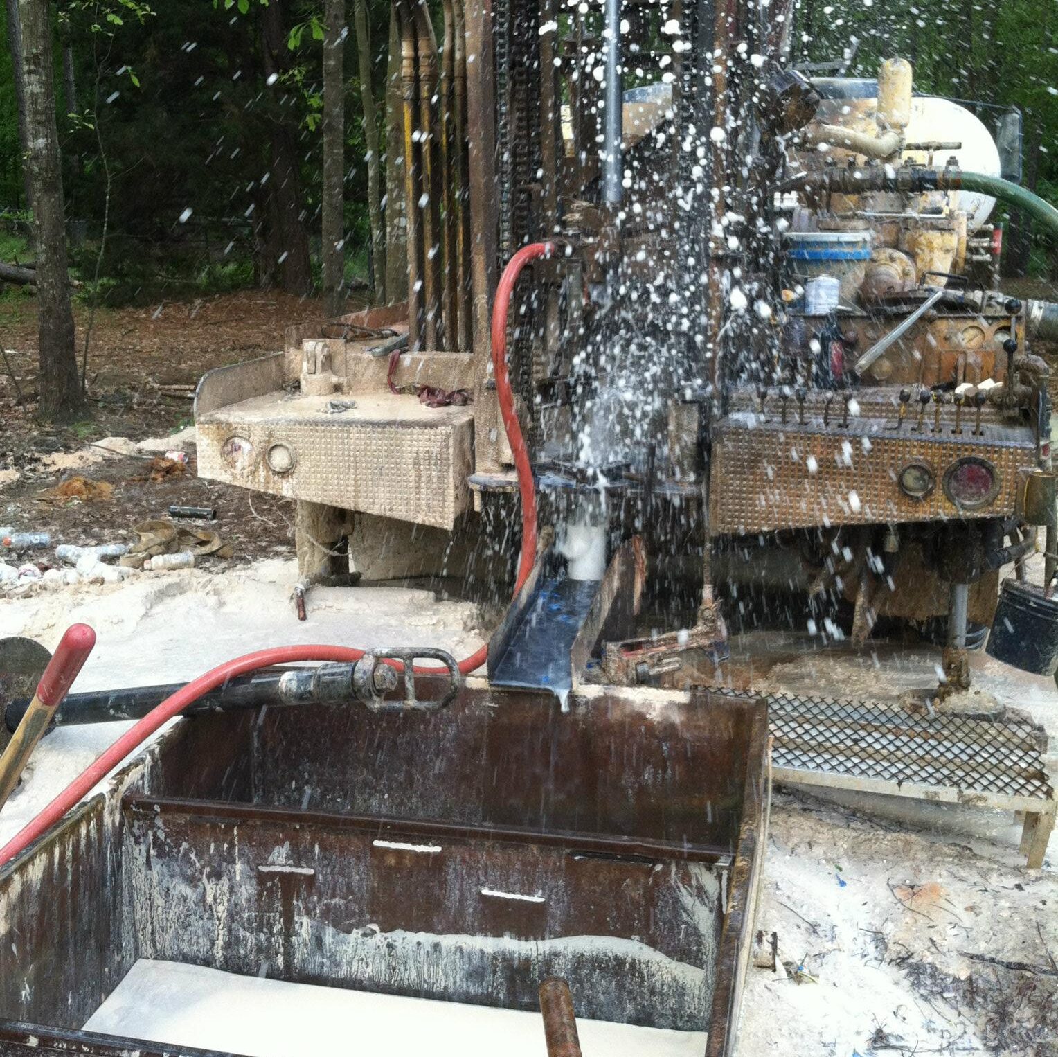 A water well drilling rig in a forested area sprays water and debris as it drills, with equipment, hoses, and containers nearby.