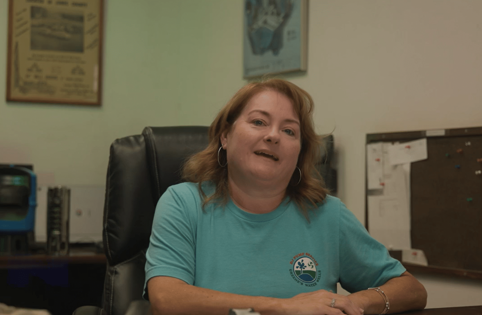 A smiling woman with shoulder-length light brown hair sits at a desk in a blue T-shirt, framed art and a corkboard behind her.