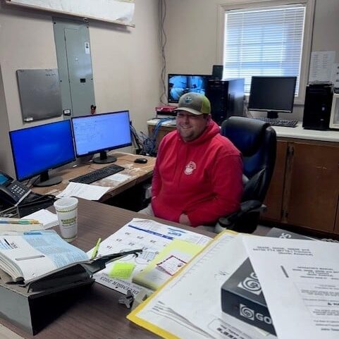 A man in a red hoodie and green cap sits at a cluttered desk with monitors, papers, office supplies, a map, window, and coffee cup.