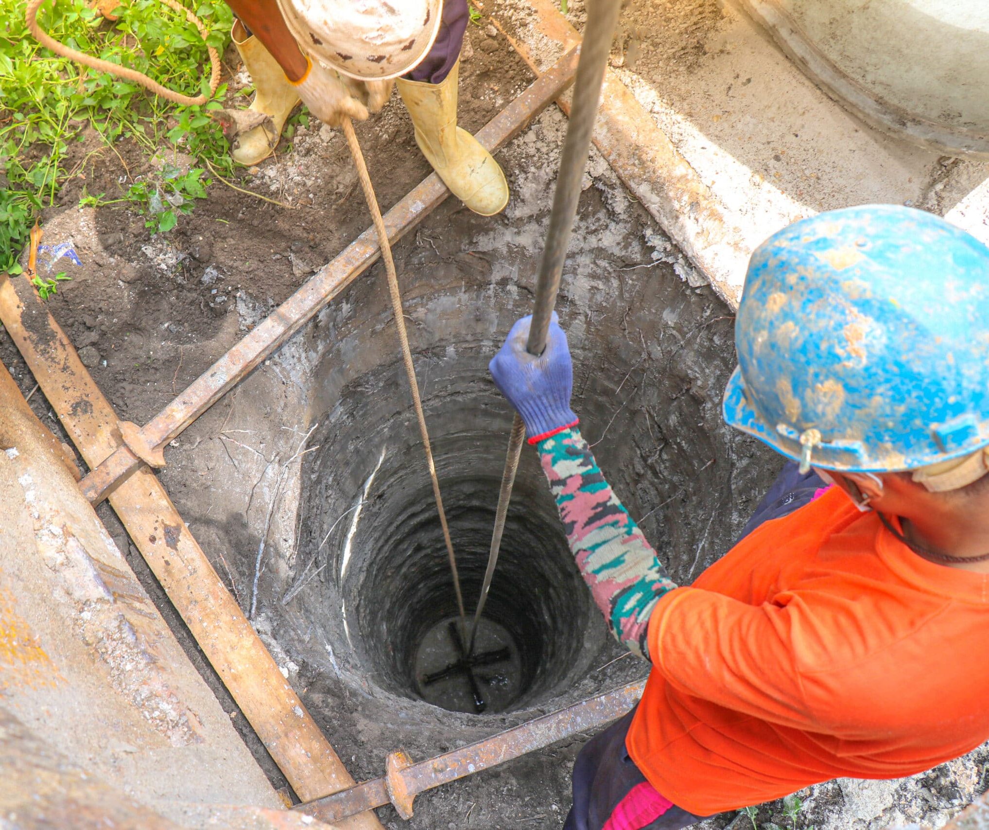 Two workers in safety gear stand by a circular well, lowering a rope or tool; metal beams and some greenery surround the area.