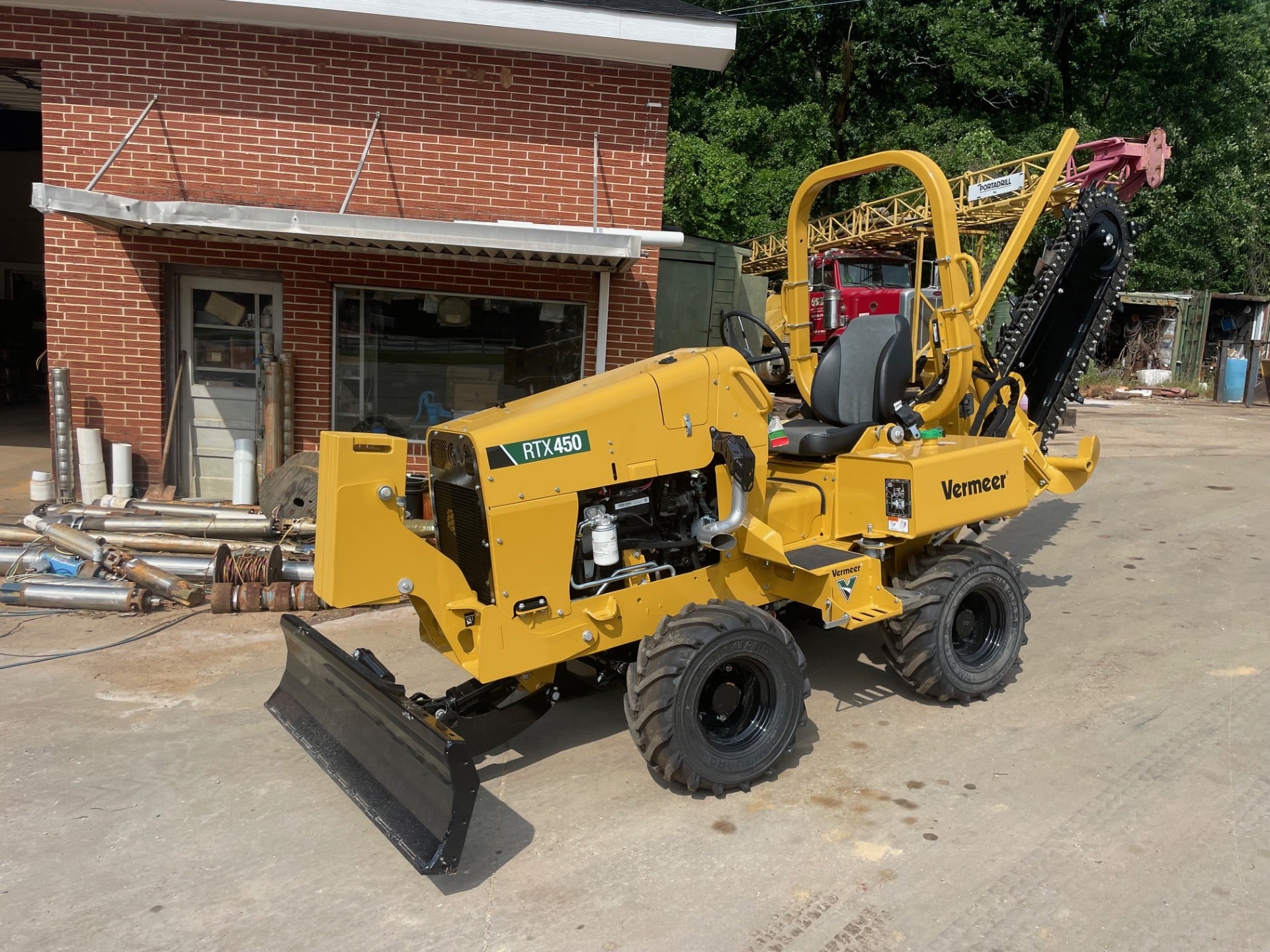 A yellow Vermeer RTX450 trencher with front blade and rear digger sits on concrete outside a brick building with large windows.