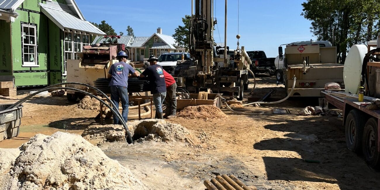 Two workers operate machinery at a construction site with a drilling rig. A green house, trees, and blue sky are in the background.