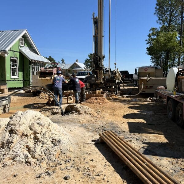 Two workers operate machinery at a construction site with a drilling rig. A green house, trees, and blue sky are in the background.