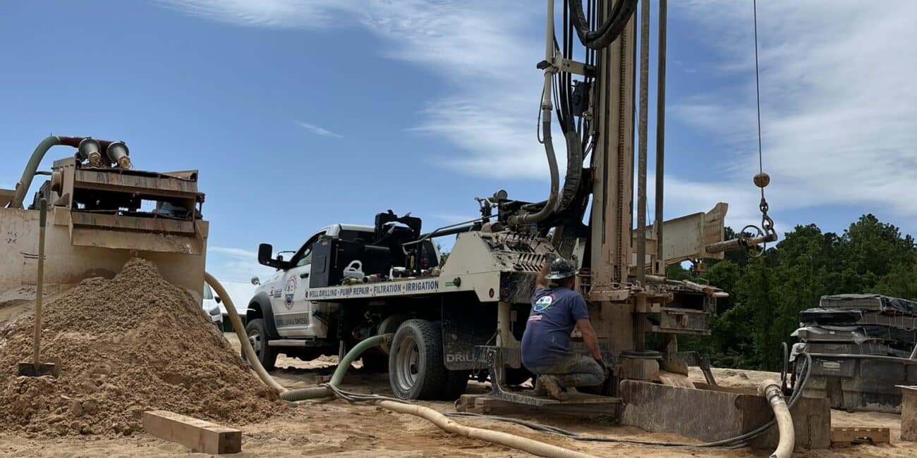 A worker kneels by a big drilling machine on a sandy site, with equipment, hoses, and sand piles under a partly cloudy sky.