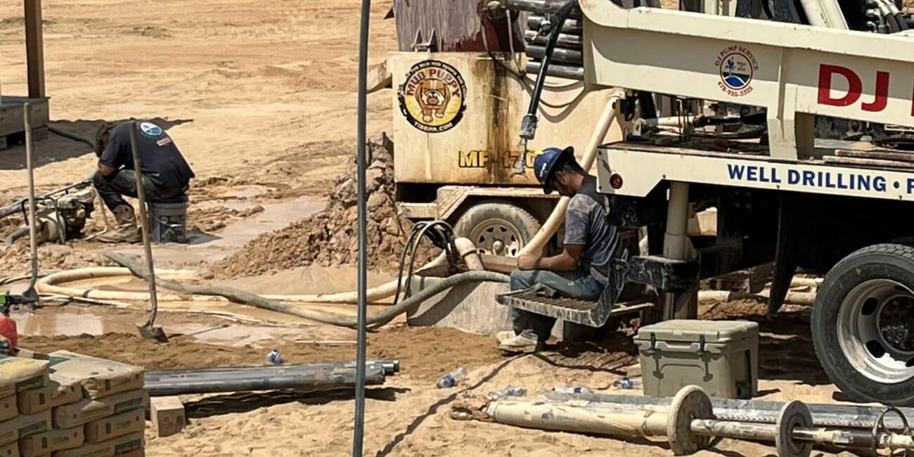 Two workers use a well drilling rig at a sandy site, surrounded by equipment and pipes, with trees in the background under clouds.
