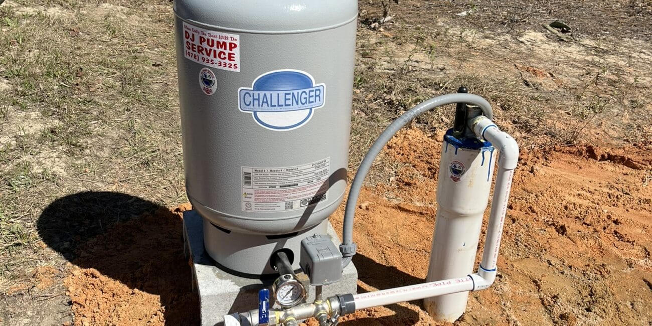 A gray Challenger pressure tank and water filtration system are installed outdoors on a concrete pad with reddish soil and trees nearby.