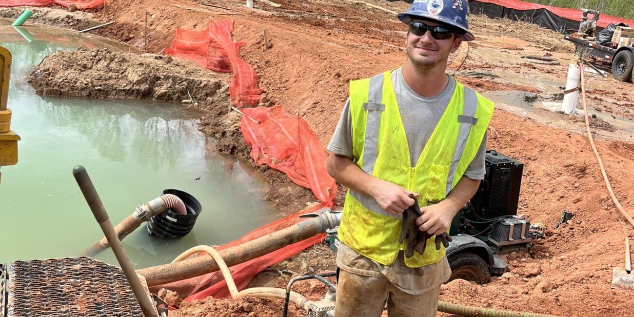 A construction worker in a blue hard hat, safety glasses, and yellow vest smiles near muddy water and equipment at a tree-lined jobsite.