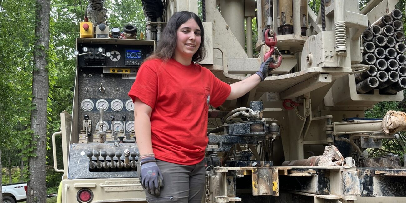 A person in a red shirt, gray pants, gloves, and boots stands on industrial machinery outdoors, smiling while holding a lever.