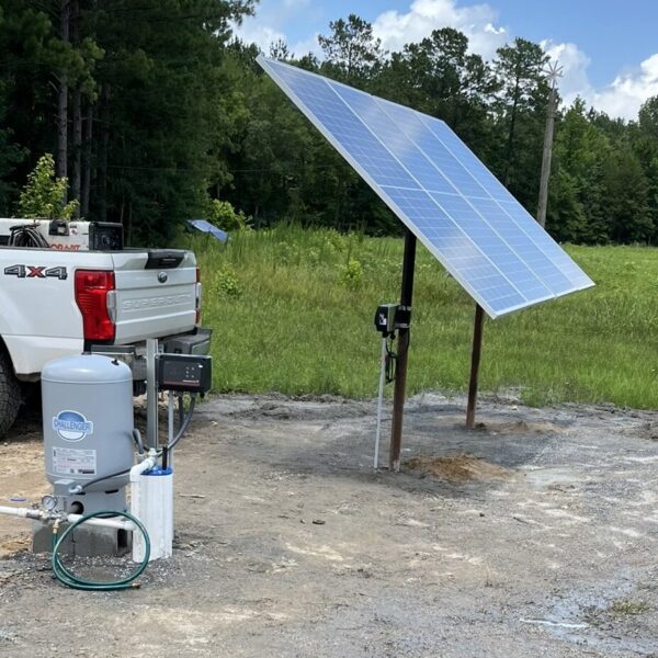 A solar panel on a metal frame stands by a white pickup truck on dirt; nearby, a pump system and blue hose sit on the ground.