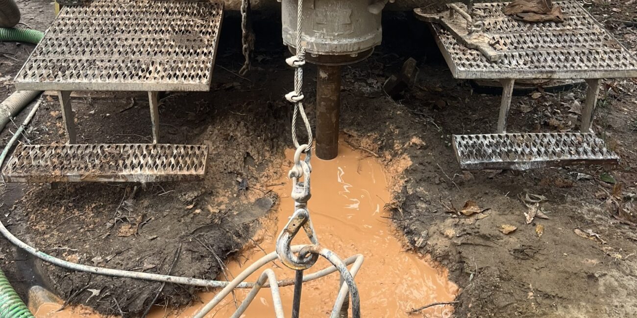 A muddy drill rig operates at a construction site, flanked by metal platforms and orange-brown water pools below. Hoses and cables attached.