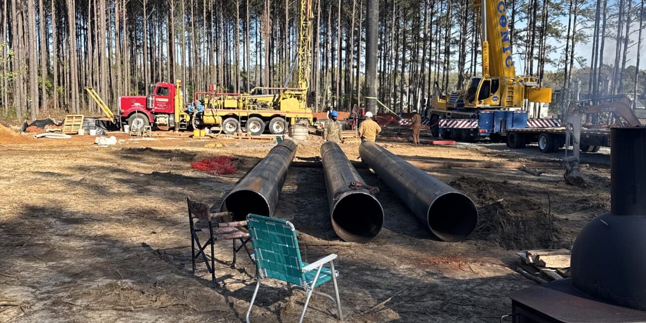 Two large yellow cranes, a red truck, and pipes at a wooded construction site. A chair and metal stove in the foreground. Pines all around.