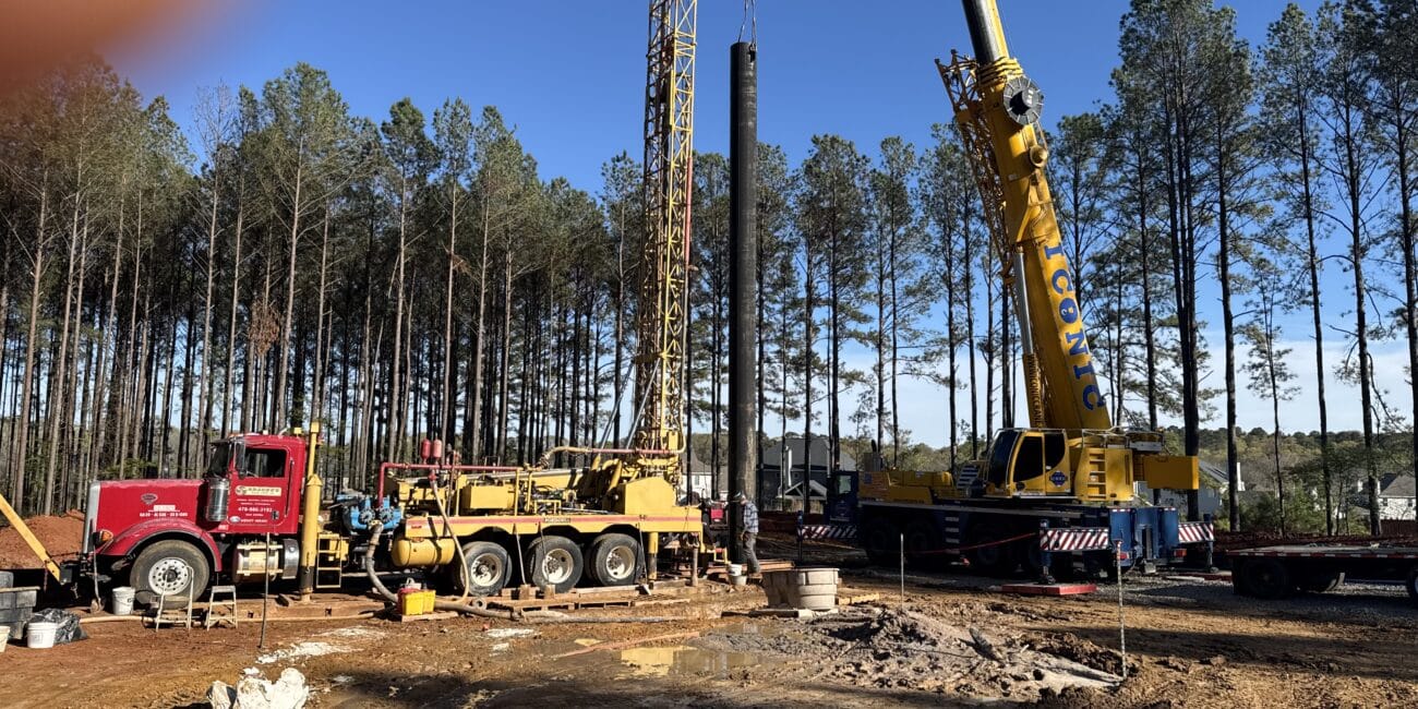 Construction site with red and yellow cranes lifting materials, tall pine trees in back, clear sky, finger partly visible top left.